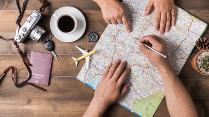 Zwei Menschen zeigen auf eine Weltkarte, Kamera, Kaffee, Reisepass und ein Flugzeug liegen auf dem Tisch
