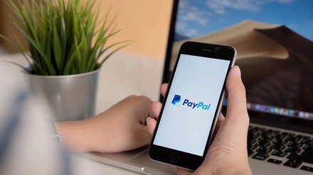 Nahaufnahme von Händen einer jungen Frau, die ein Smartphone mit eingeblendetem PayPal-Logo in der Hand hält, ein aufgeklappter Laptop steht im Hintergrund
