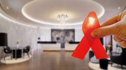 Eine Hotellobby im Hintergrund, eine Hand mit roter AIDS-Schleife im Vordergrund