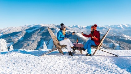 2 Frauen sitzen auf einer Skipiste in Liegestühlen und genießen ein Glas Wein