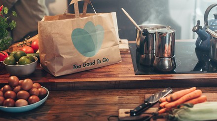 Eine „Too Good To Go“-Tüte steht auf einer Küchenplatte mit Herd, drum herum liegen verschiedene Lebensmittel