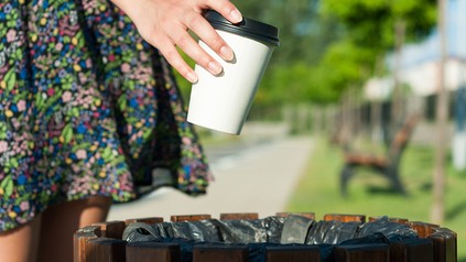 Nahaufnahme einer Frauenhand, die gerade einen Einweg-Kaffee-to-go-Becher in einen Mülleimer in einem Park wirft
