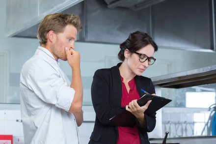 Frau notiert sich etwas auf Klemmbrett, Koch sieht besorgt zu