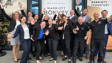 Gruppenfoto der Mitarbeiter des Marriott Bonvoy in Frankfurt, die mit ausladenden Gesten vor der Fassade des Hotels posieren