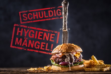 Ein Hamburger, in dem ein Messer steckt. An der Wand dahinter steht „schuldig!“