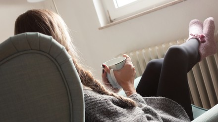 Junge Frau sitzt in einem Sessel, eine Tasse in der Hand, die Füße auf einen Heizkörper gestellt