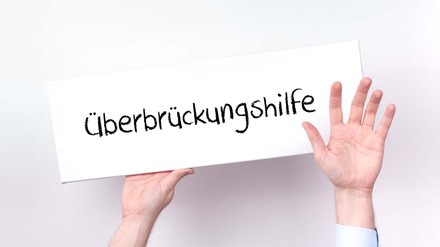 Hände halten ein Schild mit der Aufschrift „Überbrückungshilfe“ hoch