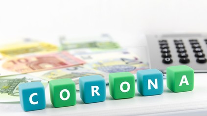 Würfel mit dem Wort Corona, im Hintergrund Euro-Geldscheine und ein Taschenrechner