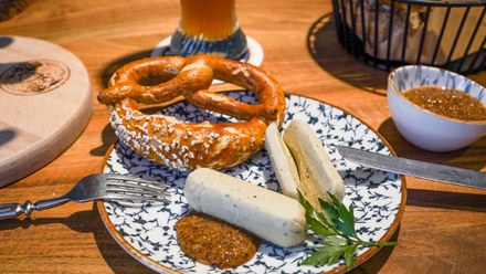 Stylischer Frühstücksteller mit Weißwurst, Brezel und süßem Senf. Im Hintergrund ein gekochtes Ei