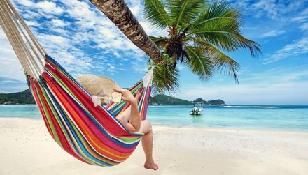 Eine Frau liegt mit einem Sonnenhut entspannt an einer Hängematte an einer Palme am Strand